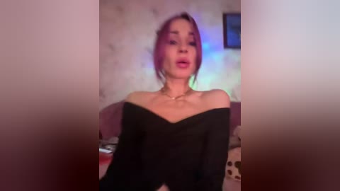 Video of a young woman with light skin, wearing a black off-shoulder top, pink lipstick, and earrings, seated on a bed with floral-patterned bedding in a dimly lit, softly colored room.