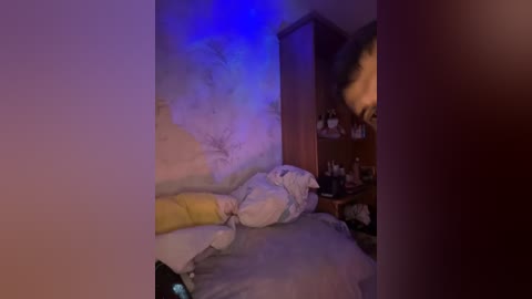 Video of a dimly lit, messy bedroom with a bed covered in white and yellow sheets, a wooden dresser with bottles and a lamp, and a blue-tinted wall.
