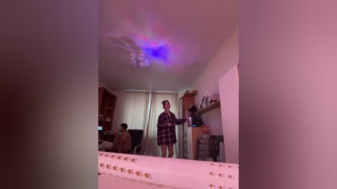 A video of a dimly lit, cluttered bedroom with a woman in a plaid nightgown, standing in front of a mirror, illuminated by a multicolored laser light.