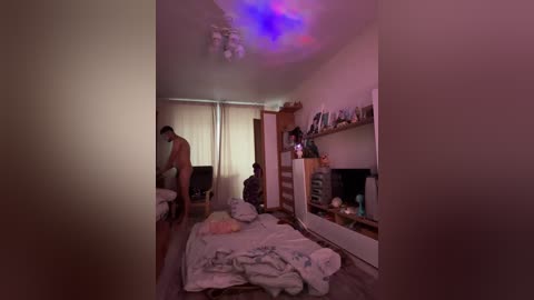 Video of a dimly lit, messy bedroom with two naked people, one standing, one sitting on a bed, under a purple LED light.