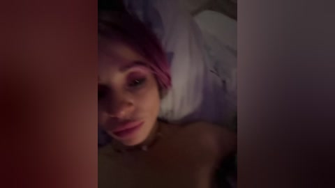 A dimly-lit video shows a close-up of a young woman with pink hair, lying on a bed with white sheets. She has fair skin and wears pink eyeshadow. The image is blurry.