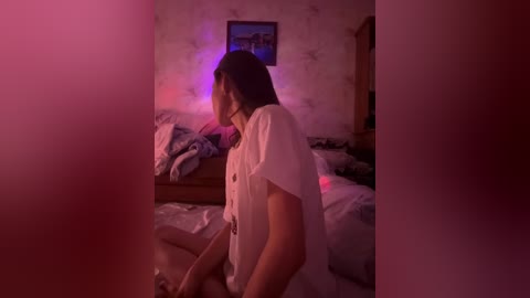 A dimly lit video of a young woman with long dark hair, wearing a white blouse, standing in a cluttered bedroom with pink and purple lighting.