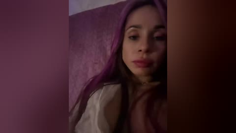 A dimly lit video of a young woman with long, purple hair, wearing a light-colored jacket, standing against a dark, textured wall.