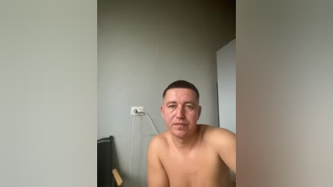 Video of a shirtless, light-skinned man with short brown hair, looking serious, in a sparsely furnished room with light gray walls, a power outlet, and a black speaker.