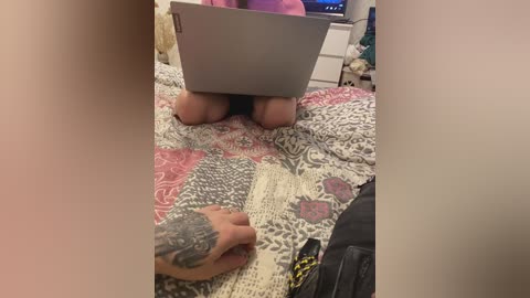 Video of a person with light skin, tattoos on their forearm, wearing a pink shirt, kneeling on a patterned quilt, working on a laptop. Background includes a nightstand with a lamp and a TV screen displaying a blue light.