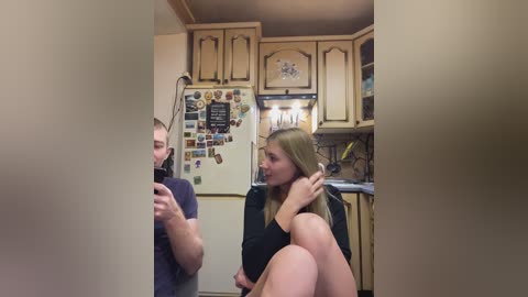 Video of a cozy kitchen with wooden cabinets, a fridge adorned with magnets, and a woman with long blonde hair sitting on the floor in a black top.