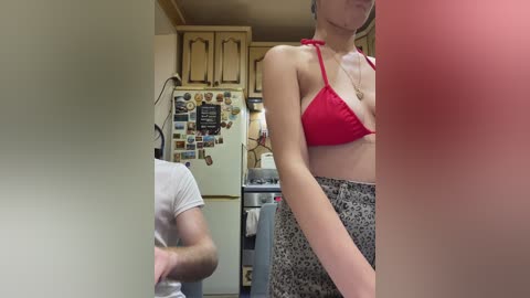 Video of a woman in a red bikini top and leopard-print skirt, standing in a cluttered kitchen with a refrigerator adorned with magnets and a man partially visible in the background.