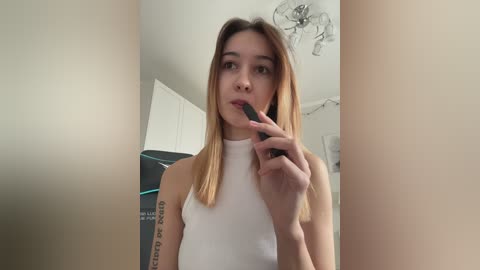 Video of a young, fair-skinned woman with long, straight blonde hair, wearing a white, sleeveless top, standing in a modern kitchen, holding a black lipstick near her lips.