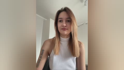 A video of a young, light-skinned woman with straight, shoulder-length blonde hair, wearing a white sleeveless top, standing in a minimalist room with beige walls and a small window.