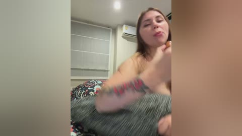 A candid photo of a topless woman with light skin, shoulder-length brown hair, and a tattoo on her arm, lying on a bed in a dimly lit room with a patterned blanket, white blinds, and an air conditioner.