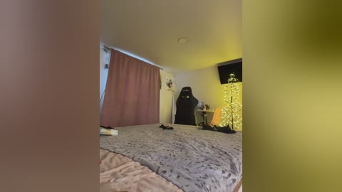 Video of a dimly lit bedroom with a beige bedspread, a brown curtain, a black chair, and a small Christmas tree with yellow lights.