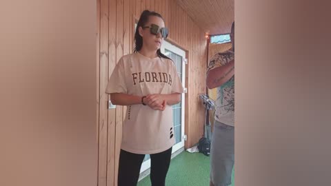 Video of a young woman with fair skin, wearing a white T-shirt with \"Florida\" and black leggings, standing in a wooden hallway with green carpet.