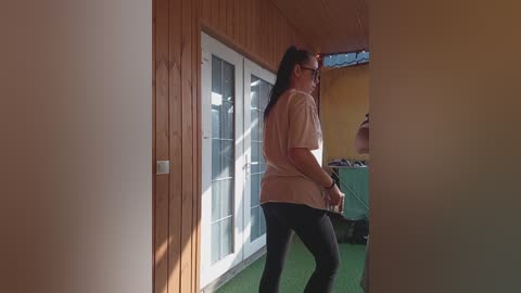 Video of a middle-aged woman with long hair, wearing glasses and a beige shirt, walking on a green carpeted balcony with wooden walls and a glass door, inside a yellow-walled room.