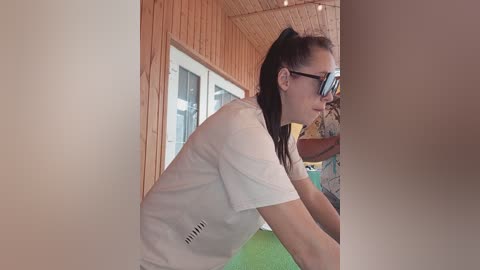 Video of a woman with black hair in a ponytail, wearing glasses and a white t-shirt, leaning over a green table in a rustic wooden room with a sloped ceiling.