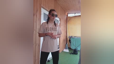 Video of a young woman with fair skin, wearing a white \"Florida\" T-shirt, black leggings, and sunglasses, standing in a narrow wooden hallway with green carpet and a turquoise laundry basket.