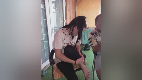 Video of a woman with long black hair, wearing a white t-shirt and black pants, sitting on a chair, holding a black hat. She's in a room with green carpet, wooden walls, and a table with a man standing.
