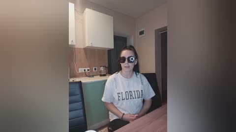 Video of a young woman with fair skin, wearing a white T-shirt with \"ELORDIA\" in bold letters, sitting at a table, wearing virtual reality goggles. The background features a modern kitchen with white cabinets and appliances.
