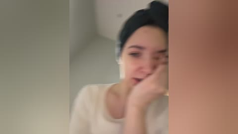 Video of a young woman with a dark headscarf, wearing a white top, leaning on a wall in a dimly lit room.