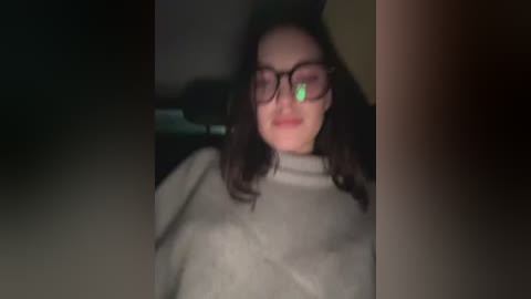 A dimly-lit video shows a young woman with shoulder-length dark hair, wearing glasses, a grey turtleneck sweater, and a slight smile. The background is blurry, suggesting an indoor setting.