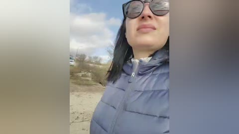 Video of a woman with long black hair, wearing sunglasses, a puffy blue jacket, and a white shirt, standing outdoors with a cloudy sky in the background.