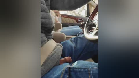 Video of a person in a car, wearing blue jeans and a dark sweater, legs resting on the passenger seat. The interior is dimly lit with beige and brown leather upholstery.