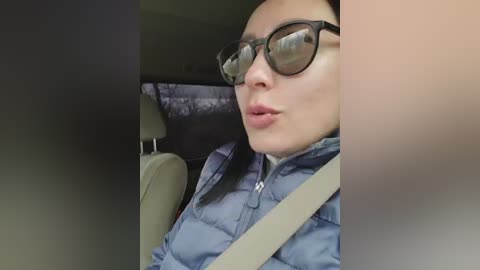 A video of a person with short black hair, wearing glasses and a blue puffer jacket, seated in a car with beige seats and a gray seatbelt.