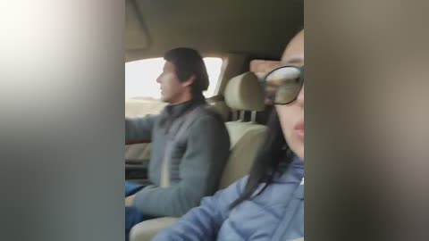 Video of a man and woman in a car, driving. The man, with dark hair, wears a gray jacket, and the woman, with long black hair, wears glasses and a blue jacket.