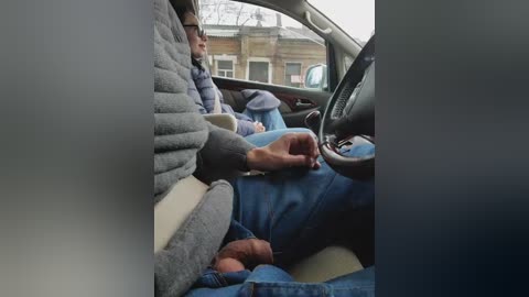 Video of a man in a gray jacket and blue jeans reclining in a car seat, with a visible erection, surrounded by urban buildings.