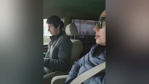 A video of a man and a woman in a car, both wearing sunglasses, the woman in a blue jacket, driving.