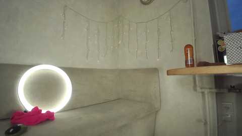 Video of a minimalist, modern interior with a circular, bright white lightbulb on a gray couch, a wooden table holding an orange bottle, and a string of white lights hanging on the beige wall.