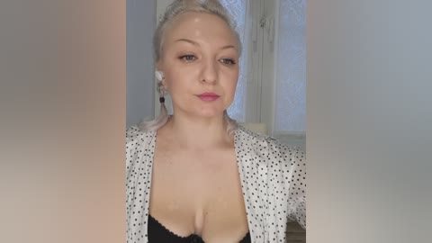 Video of a light-skinned woman with blonde hair, wearing a white polka-dot blouse and black bra, standing indoors.