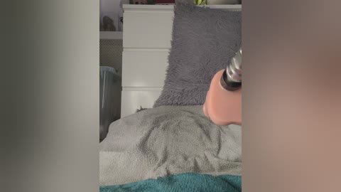 A video of a bed with a pink lamp, grey and teal blankets, and a grey pillow on a white dresser in a dimly lit room.