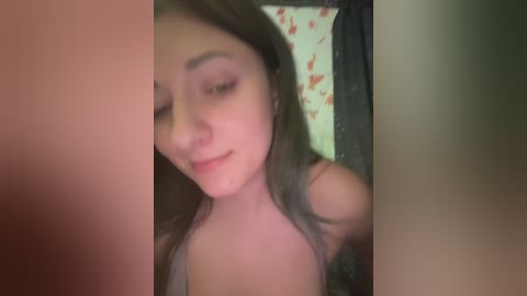 A blurry video of a young woman with long brown hair, wearing a sleeveless top, smiling slightly. The background shows a floral-patterned wall and a dark-colored door.