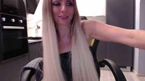 Video of a blonde woman with long, straight hair, wearing a black top, smiling, seated at a modern kitchen counter.