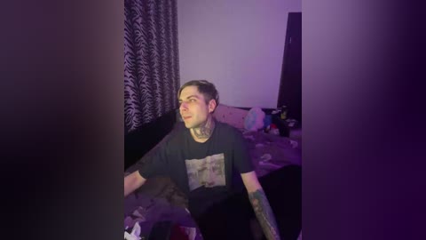 Video of a tattooed man with short brown hair wearing a black T-shirt, sitting on a bed in a dimly lit room with purple lighting.