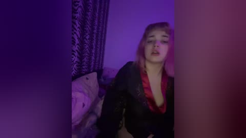 Video of a young woman with blonde hair, wearing a black robe, partially open to reveal a red bra, in a dimly lit bedroom with purple and blue lighting.