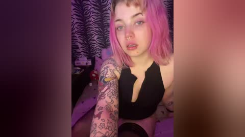 Video of a young woman with pink hair and multiple tattoos, wearing a black halter top, sitting on a bed with a zebra-patterned curtain in the background.