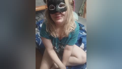 Video of a fair-skinned woman with long blonde hair, wearing a black mask with white designs and a teal T-shirt, sitting on a bed with blue sheets, in a dimly lit room.
