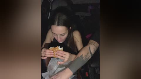 A candid video of a young woman with long dark hair, wearing a black top, eating a sandwich. She has tattoos on her arm. The background is dimly lit, showing a dark room with indistinct objects.