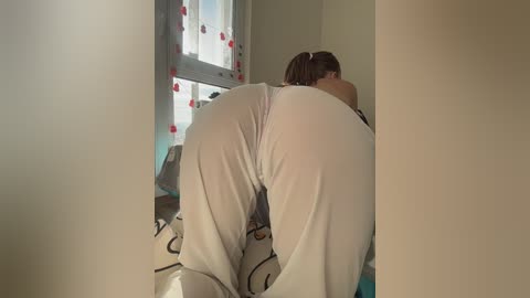 Video of a woman with light skin, long brown hair, and a curvy figure, wearing white leggings, kneeling on a bed, facing a window with red flowered curtains.