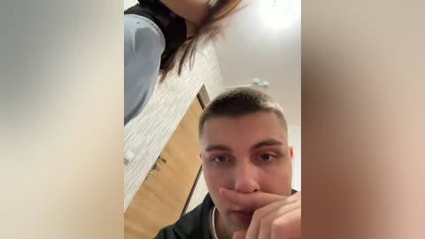 Video of a man with short dark hair, wearing a black jacket, standing in a bathroom with white tiled walls and a wooden door, looking down at a woman with long dark hair.