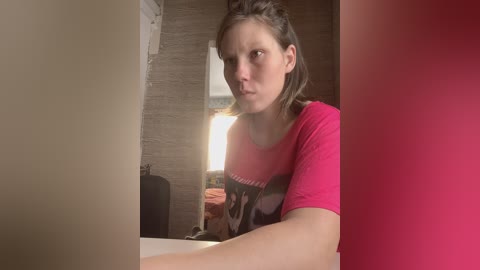 Video of a young woman with shoulder-length brown hair, wearing a pink T-shirt with a cartoon character, standing in a small, dimly lit bathroom with beige and pink walls.