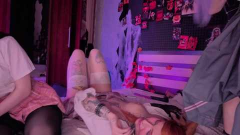 Video of a young woman with pale skin, red hair, and glasses, lying on a bed in a dimly lit room with posters and red curtains. She wears a white shirt and black stockings.