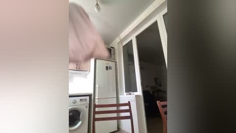 Video of a small, bright kitchen with white appliances, a wooden chair, and a blurry person in motion. The background shows an open doorway leading to a dimly lit living area.