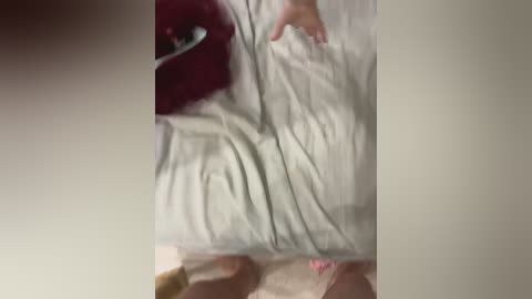 A blurry video showing a person with light skin and long hair, partially covered by a white sheet, lying on a beige carpet. The image has a dim, indoor lighting with a slight red tint.