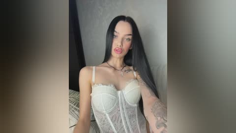 Video of a slender, fair-skinned woman with long black hair, wearing a white lace corset, standing in a dimly lit room with textured walls.