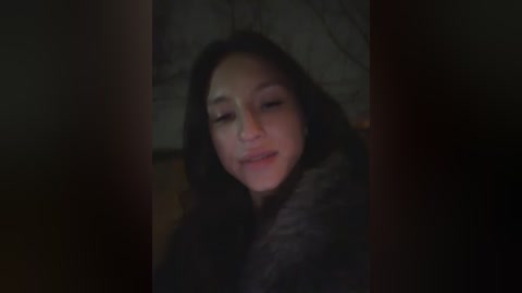 Video of a young woman with long, dark hair, lying down in a dimly lit room. She has a serene expression, eyes closed, wearing a dark fur coat. The background is blurry, with indistinct shapes.