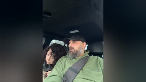 A video of a man with a gray beard and cap driving, with a woman wearing a hat in the passenger seat.