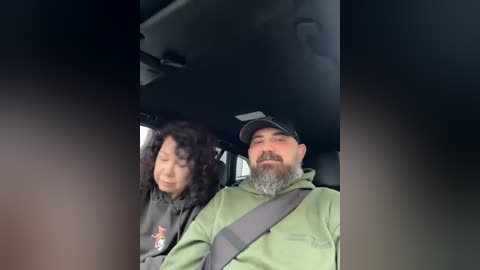 Video of a man with a beard and mustache wearing a green hoodie, driving a car with a woman beside him.