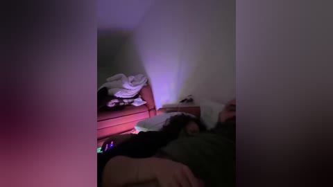 A dimly lit video shows a person lying on a bed, covered in a green blanket, with a white sheet and pillow. The room has a purple light, creating a moody atmosphere.